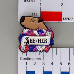 NerdyKeppie Capybara Enamel Genderfluid Pride Pronoun Pins. Our enamel pin features an adorable capybara, a paper scroll with three different pronoun choices, and flowers in genderfluid pride colors. This pride pin ships from Portland, Oregon.