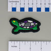 NerdyKeppie's Painted Turtle Embroidered Pride Patch are shaped like their namesake and stitched on black duck cloth!