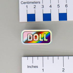 NerdyKeppie "DOLL" Mirrored Pride Pin. Mirrored pride pin with "DOLL" text in your choice of Progress, rainbow, or transgender pride colors. Say it with your whole chest.